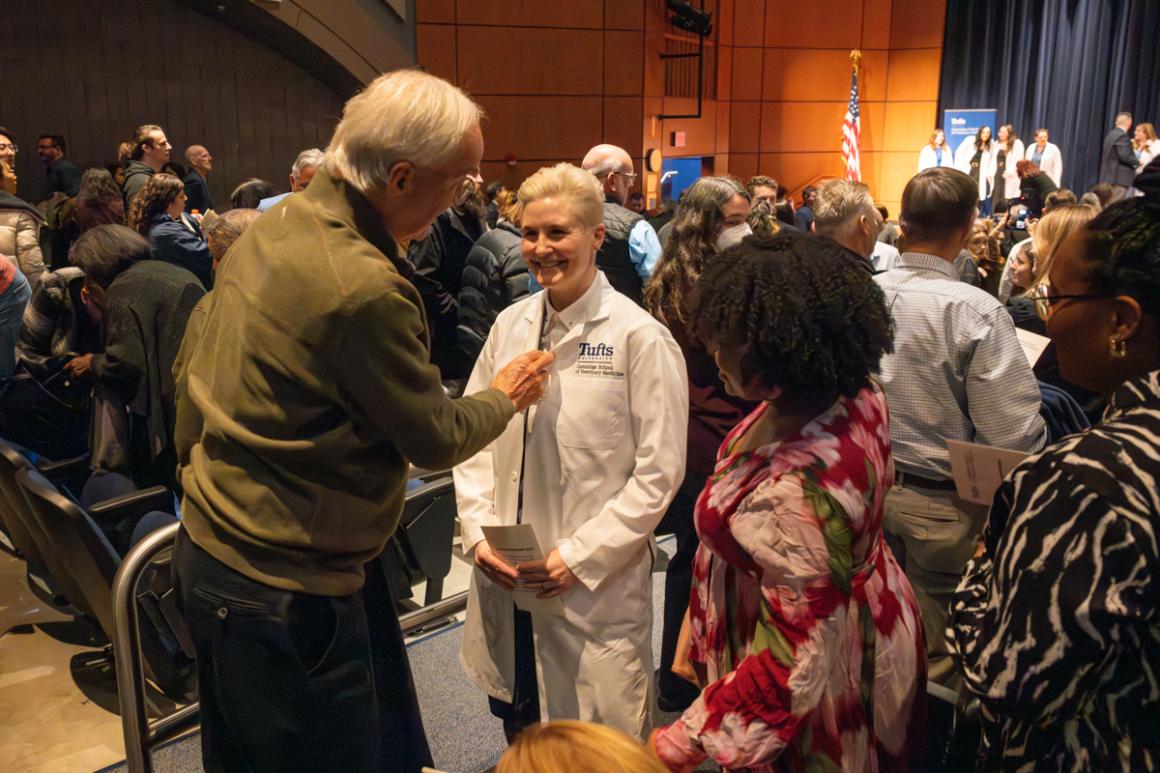 A standing older individual congratulates a smiling individual wearing a medical white coat..