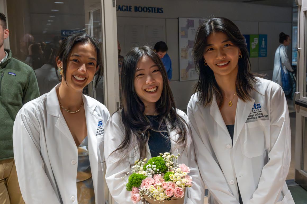 Three smiling individuals in medical white coats, one holding flowers, stand together. 