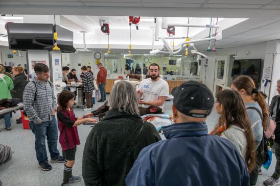An individual with a beard, wearing glasses and a white t-shirt presents to a group of visitors in a veterinary simulation lab..