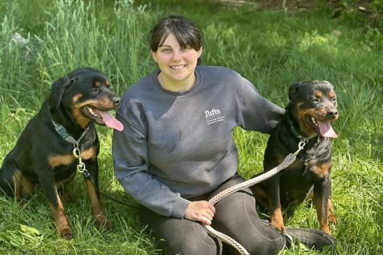 A person wearing gray pants and a gray top in the green grass with two black and brown dogs with their pink tongues out.