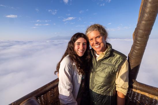 Two individuals  standing close to one another smiling with clouds in the background.