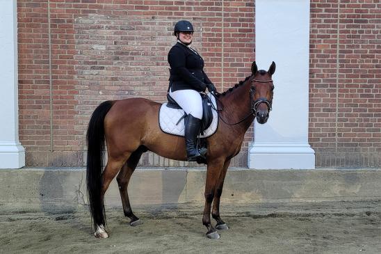 A smiling rider wearing a black helmet, black jacket, white breeches, and tall boots sits on a brown horse with a braided mane, standing on sandy ground in front of a red brick wall with white columns.