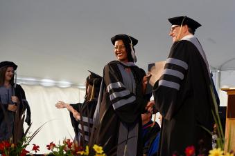 Black female graduate student shaking hands with the Dean while receiving her diploma. Both are wearing caps and gowns.