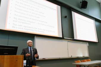 an older man with grey hair wearing a suit and tie is lecturing in a lecture hall with 2 screens behind him.