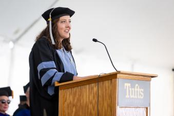 A smiling woman with long curly brown hair, wearing a cap and gown, speaking behind a podium at a commencement ceremony.