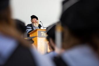 A man wearing black reading glasses, has a long beard, wearing a cap and gown, stands behind a podium addressing a graduating class of students.