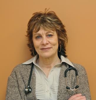 An individual with wavy brown hair and blue eyes wearing a collared shirt, a sweater, and a stethoscope