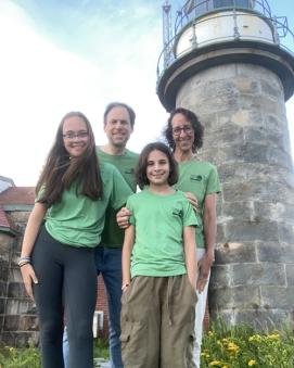A smiling family of four: mom, dad and two daughters standing in front of a lighthouse.