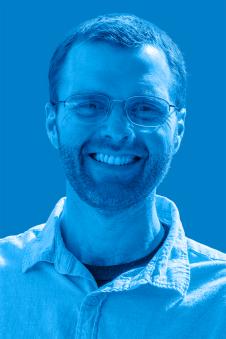  John Bourgeois smiling with short hair and facial hair wearing glasses with a tinted blue background