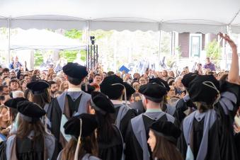 A diverse group of people, including graduates dressed in their regalia, stands under a large tent outdoors at a commencement ceremony.