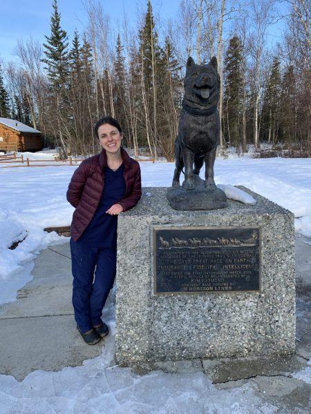 Women standing next to the Iditarod Race monument with a statue of a sled dog on top