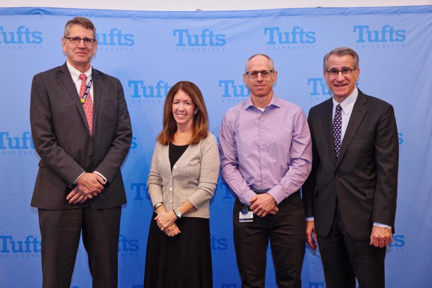 Three men and one women posing for a group photo with a Tufts blue banner in the background