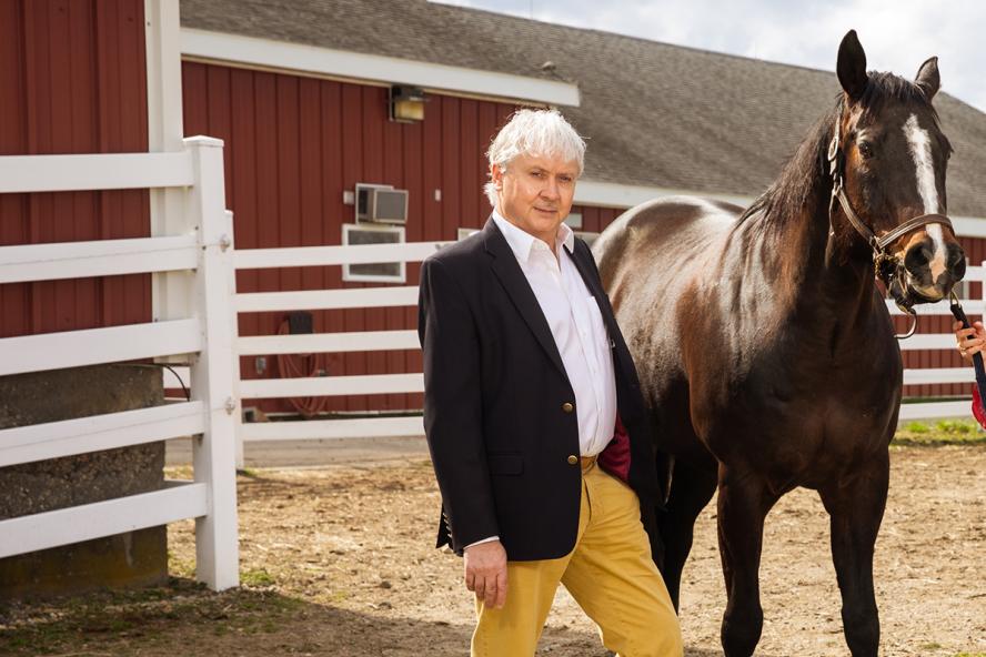 an Englishman with grey hair standing next to a horse in front of a barn. He's wearing a white dress shirt and black blazer.