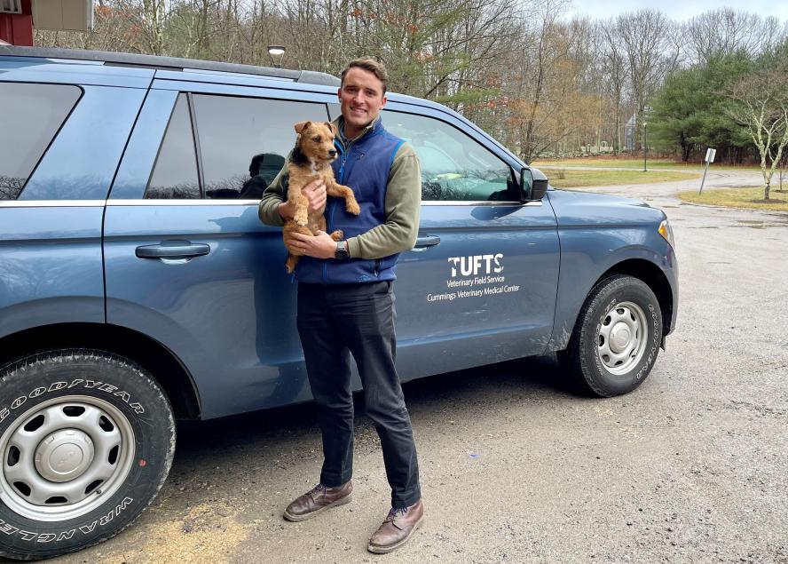 A man wearing a blue vest holding his small dog standing in front of a blue SUV with the Tufts logo on the side door