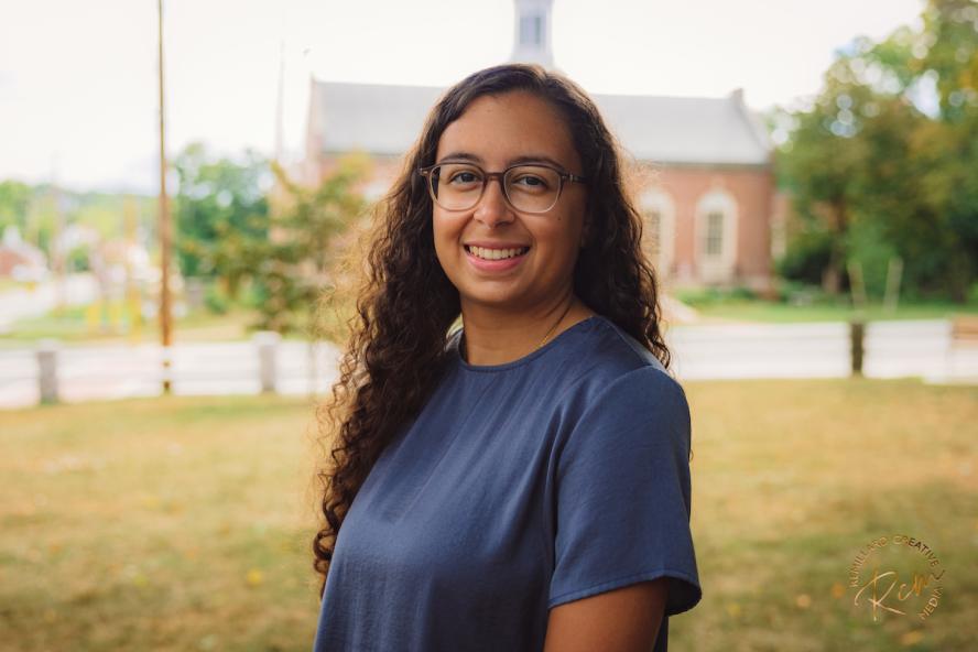 A woman with long, curly brown hair, wearing glasses and a blue T-shirt smiles while having her photo taken outdoors.