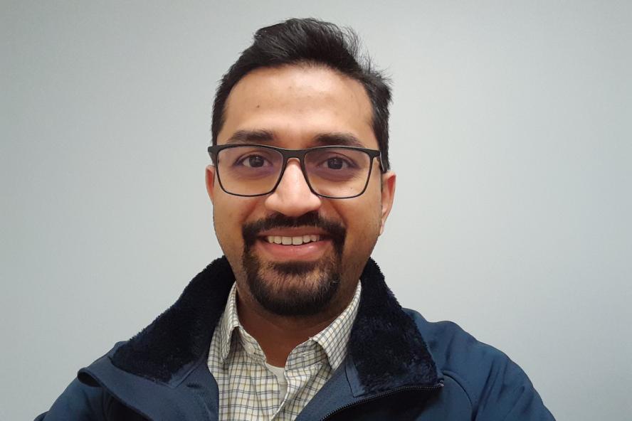 A smiling man from India with a circle beard with soul patch and black hair is wearing dark-rimmed glasses and a button-down collared shirt under a navy zippered coat.