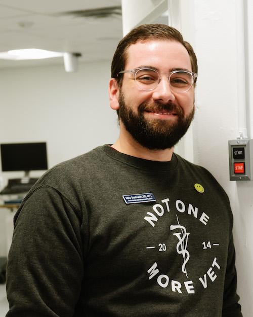A smiling man with brown hair, beard and mustache wearing glasses and a long sleeve shirt that says 'Not one more vet'