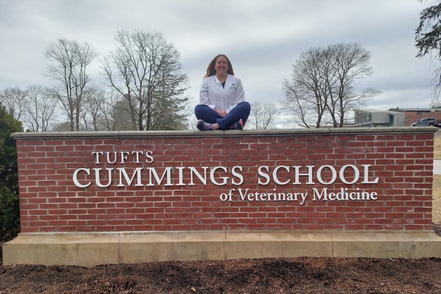 A woman wearing a white coat and blue jeans sits atop a brick wall with sign attached that reads Tufts Cummings School of Veterinary Medicine.