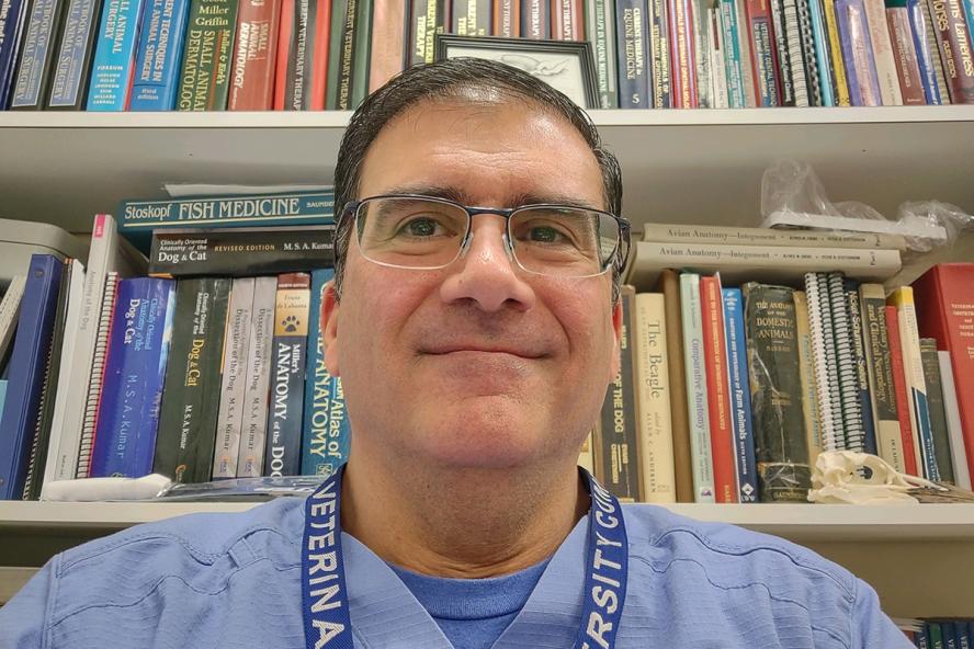 man wearing glasses and blue hospital scrub smiling and stading in front of shelves of books