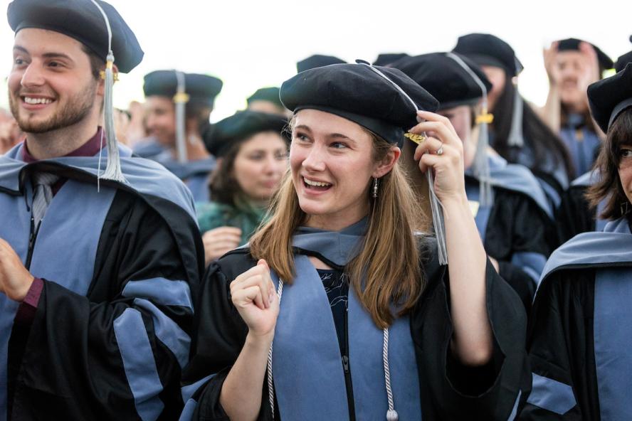 Cummings School doctor of veterinary medicine graduates wearing blue and black gowns and black barets. A smiling man in front is clapping and a smiling woman with long light brown hair is moving her tassel to the other side of her cap.
