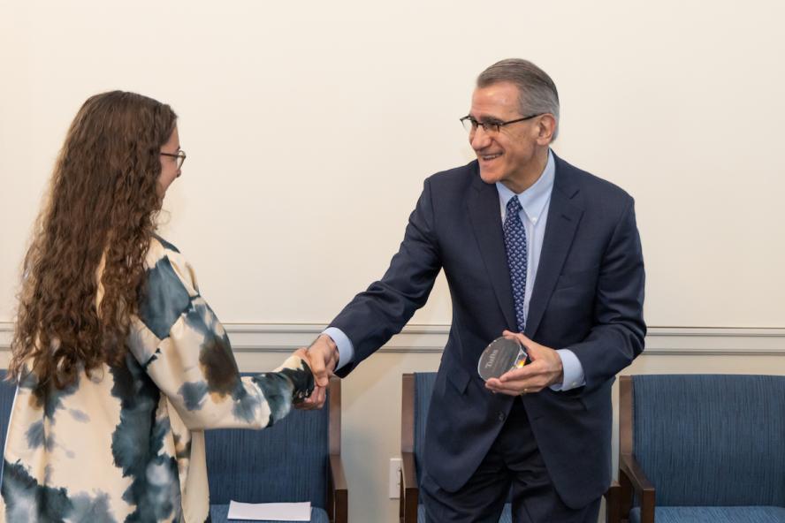 Woman with long curly brown hair receiving an award from a smiling man wearing glasses, suit and tie. They are shaking hands.