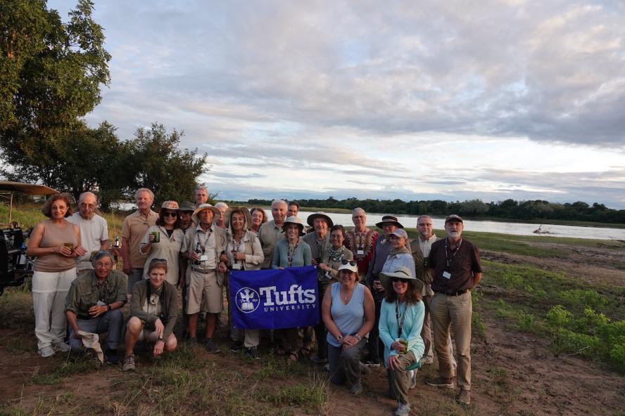 A group of 24 people dressed in neutral colored clothing posing for a photo in South Luangwa Zambia. They are holding a blue Tufts sign representing the Tufts Travel-Learn participants.