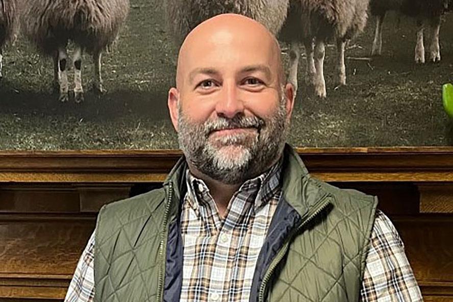 A smiling man with a beard and mustache, wearing a green vest and plaid shirt.