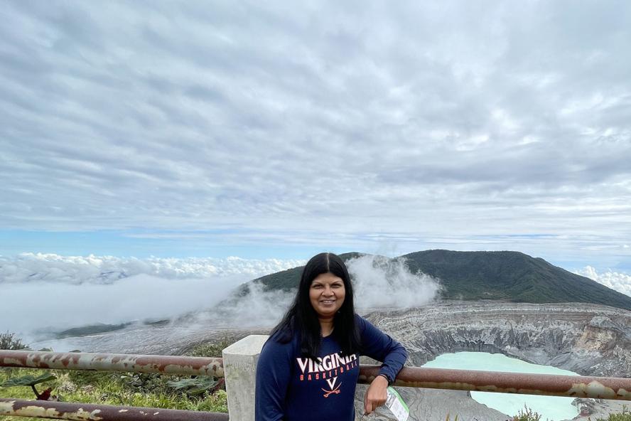 A smiling woman with long black hair, wearing a long sleeve navy sweatshirt, is posing for a photo while leaning on a railing in front of a volcano and a crater lake filled with light blue water.