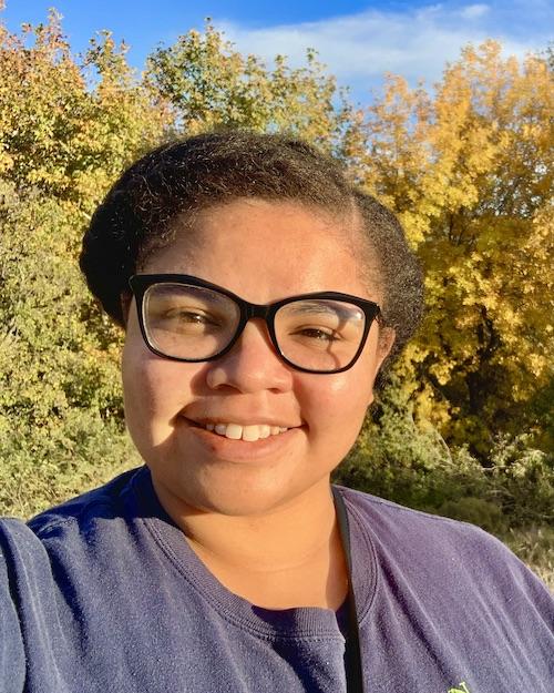 A smiling woman of color with short hair wearing glasses and a blue sweater. Trees and foliage in the background.