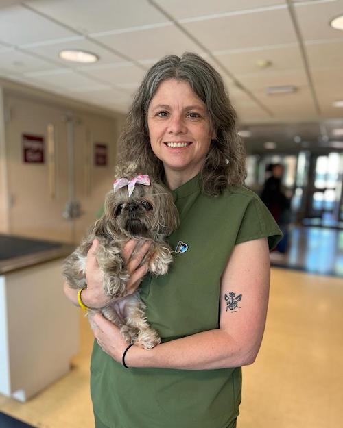 a smiling woman with grey brown hair wearing a dark green shirt and holding a little dog who has a pink bow on her head.