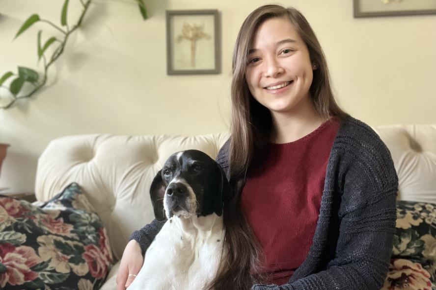 A woman with long brown hair wearing a red shirt and gray sweater sits on a couch with dog with a black and white colored dog