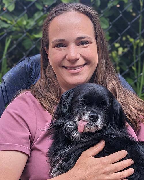 A smiling woman with long light brown hair, wearing a mauve short sleeve shirt and holding a little black dog whose tongue is hanging out.