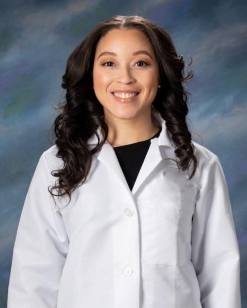A smiling woman with long dark brown curly hair wearing a white medical coat.
