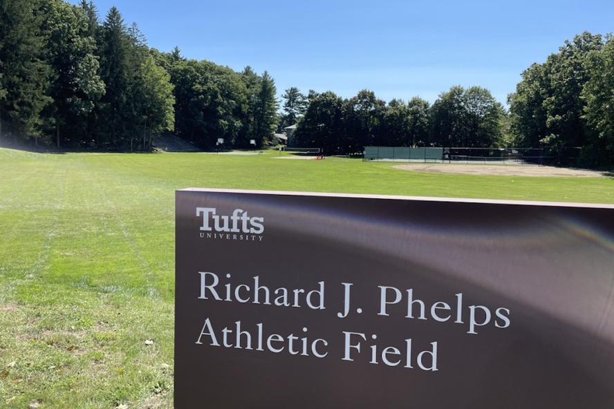 field of grass, tall bushes and trees, and a Tufts sign that reads Richard J. Phelps Athletic Field.