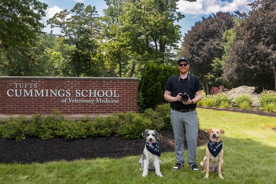 An individual wearing a baseball cap, blue polo shirt and gray pants holds a camera, flanked by a dog sitting on either side