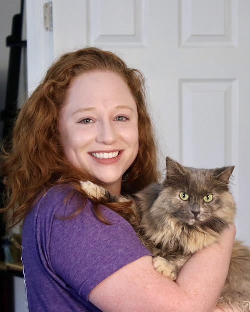 A smiling woman with long wavy red hair holding a red and brown cat.