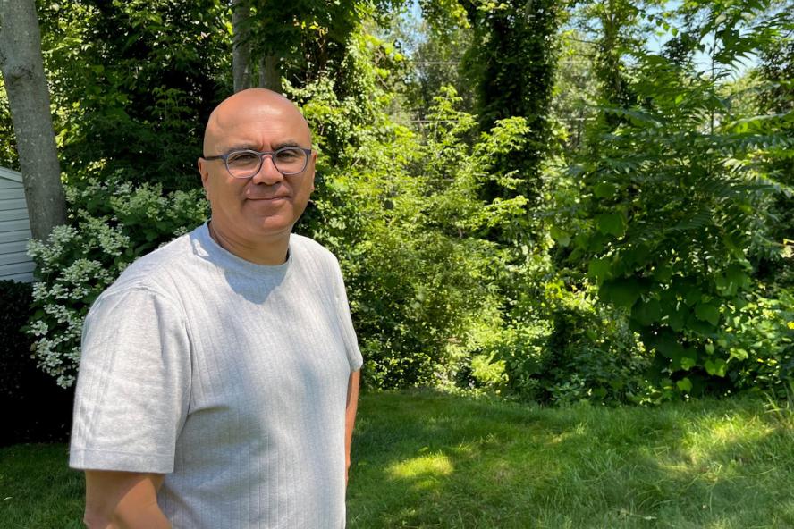 A bald man wearing glasses and a white short sleeve tee standing outdoors with trees and bushes behind him.