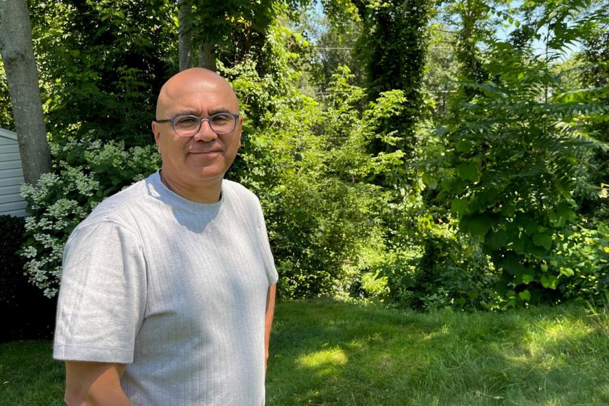 A bald man wearing glasses and a white short sleeve tee standing outdoors with trees and bushes behind him.