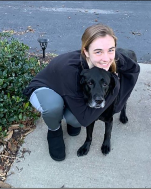 A smiling person presenting as a female is hugging her black dog.