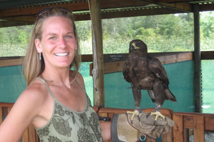 a smiling woman with long blond hair wearing a tank top. She has a steppe eagle perched on her arm.