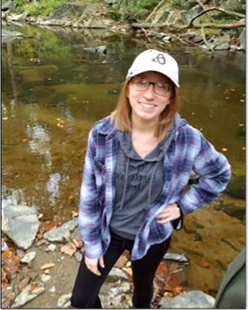 A smiling person wearing a baseball cap, glasses, blue plaid long sleeve shirt standing near a lake