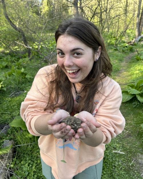a happy person with long brown hair stands in the woods holding a frog