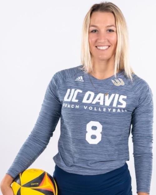 a smiling person with long blond hair wearing a UC Davis long sleeve blue shirt and holding a yellow volley ball