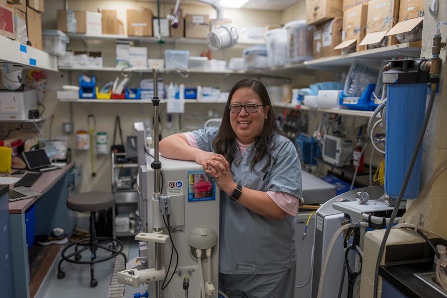 a smiling individual with long black hair wearing glasses and blue hospital scrubs