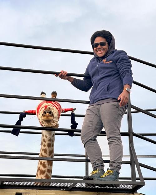 a smiling person with dark skin wearing sunglasses and a blue hoodie sweatshirt is standing on a platform holding onto a railing with a giraffe behind him