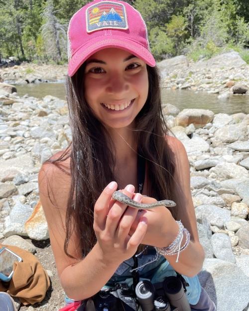 a smiling person with long dark hair wearing a pink cap is holding a small snake in an area covered with rocks and streams of water