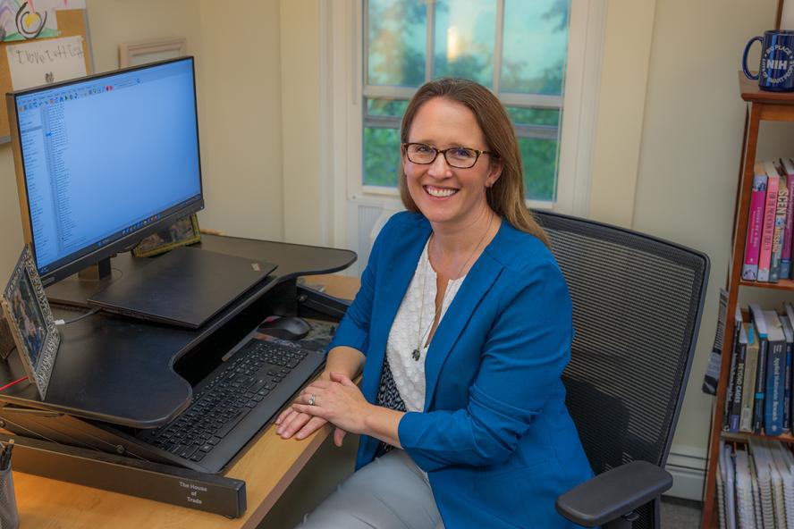 a smiling individual with long light brown hair wearing eye glasses and a blue sportcoat sits in front a computer