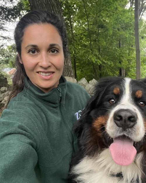 a smiling person with dark hair pulled back, wearing a green jacket and standing with her Bernese Mountain dog