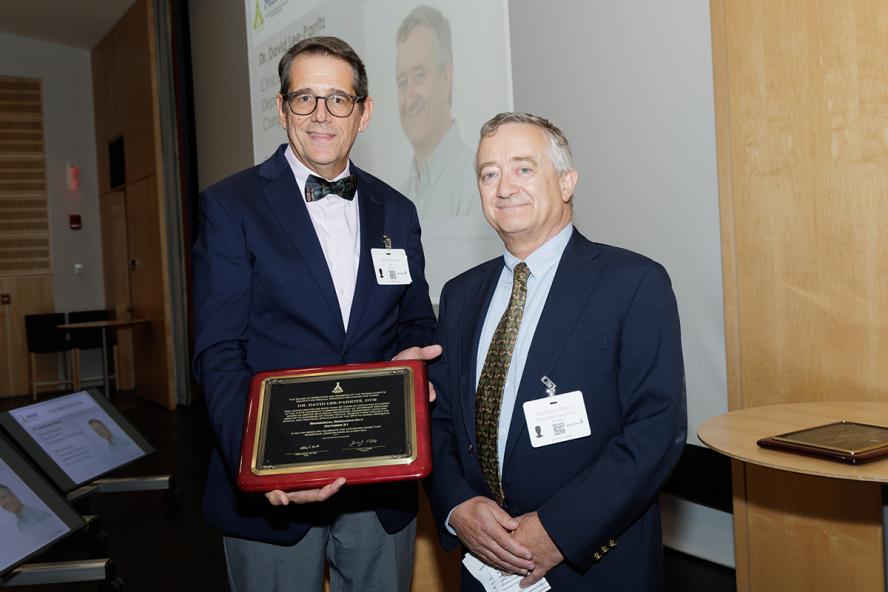 An individual wearing glasses, a navy blue suit coat, and a bowtie presents a plaque to an individual wearing a navy blue suit coat and necktie.