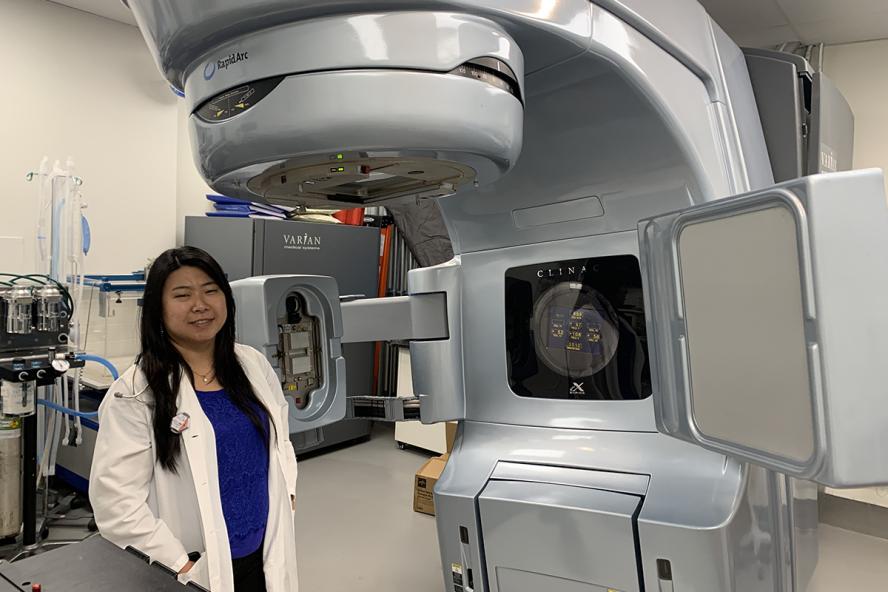 an individual with long black hair wearing a white lab coat stands beside a linear accelerator